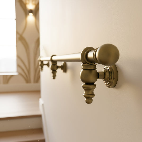 Aged brass finish stair handrail with round steel rail, ball end cap and ornate wall brackets beside a staircase